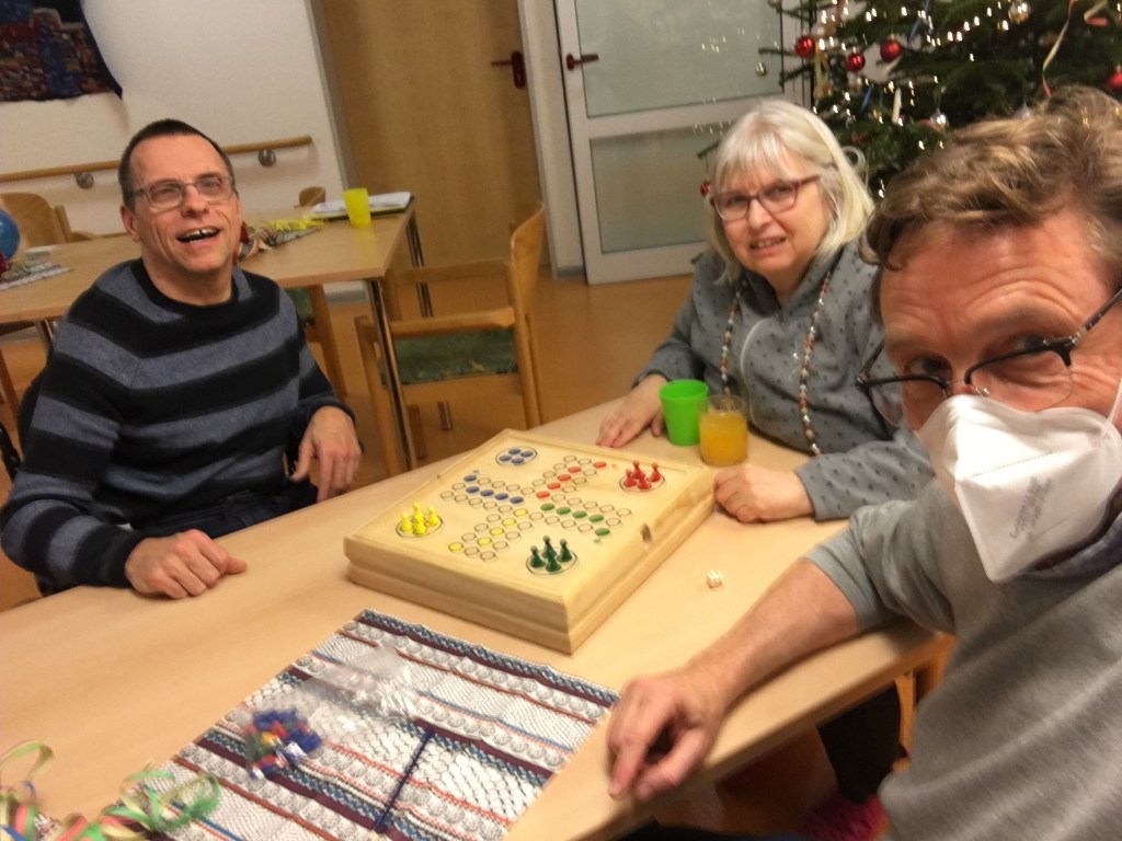 Marc van der Meer wearing a mask, sitting with a disabled man and a woman, smiling while playing a board game together at a table, with festive decorations in the background.