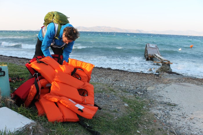Matc van der Meer wearing a blue jacket and green backpack is organizing orange life jackets on a beach in Greece with waves in the background.
